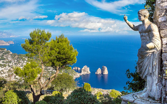 Capri coastline view with Faraglioni rocks and statue, seen from a hillside vantage point.