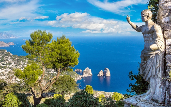 Capri coastline view with Faraglioni rocks and statue, seen from a hillside vantage point.