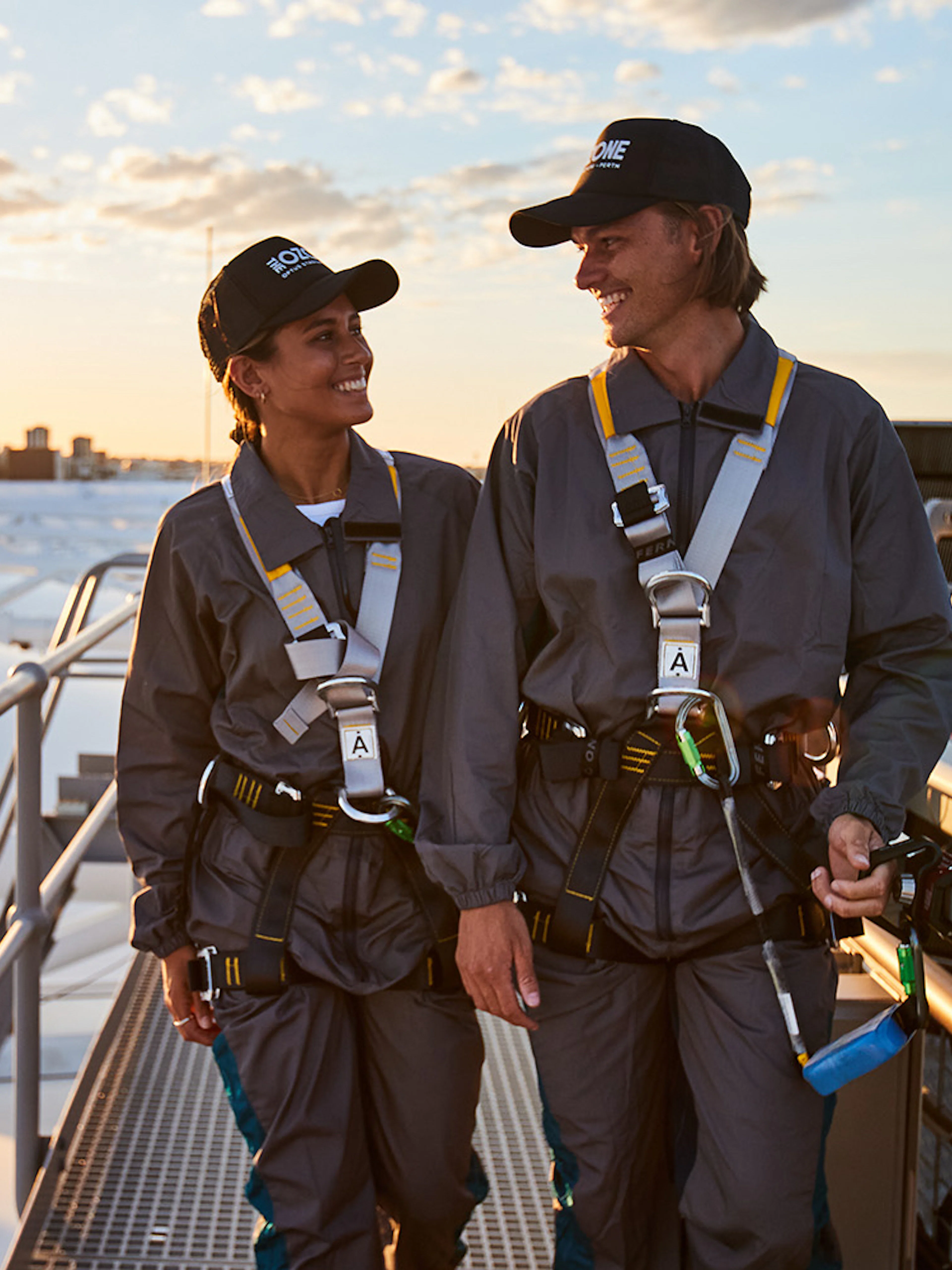 Two people in safety gear walking on Optus Stadium roof with Perth skyline in background.