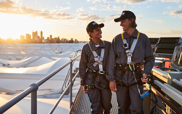 Two people in safety gear walking on Optus Stadium roof with Perth skyline in background.