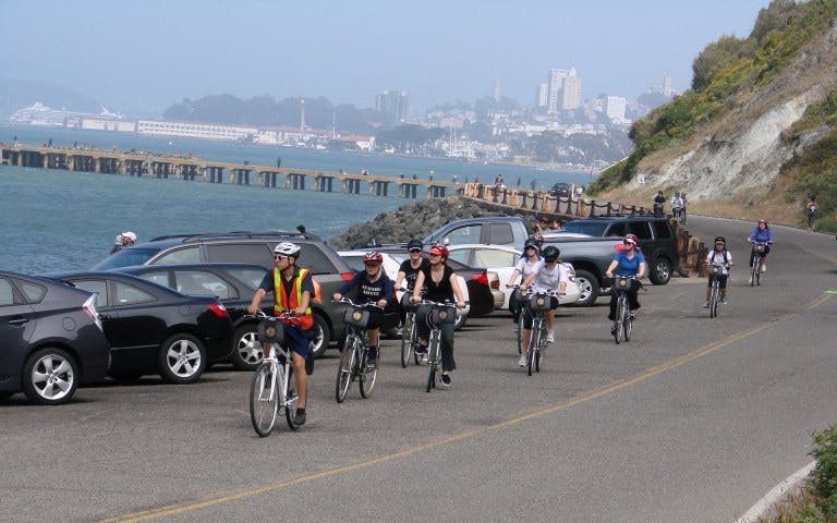 Cyclists on a coastal road during a sunset bike tour with city skyline in the background.