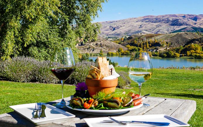 Gourmet platter and wine glasses on a table overlooking a scenic lake near Queenstown.