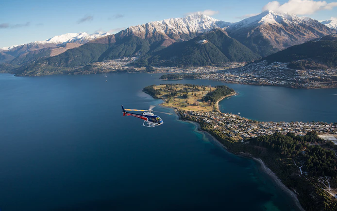 Helicopter flying over Lake Wakatipu with Queenstown and snow-capped mountains in view.