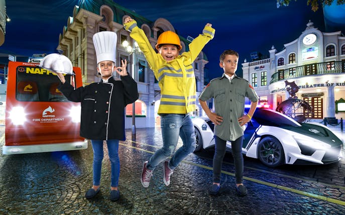 Children in firefighter, chef, and pilot costumes at KidZania Dubai with fire truck and police car.