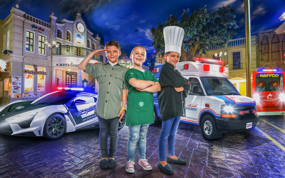 Children in role-play costumes at KidZania Dubai with police car and ambulance in background.