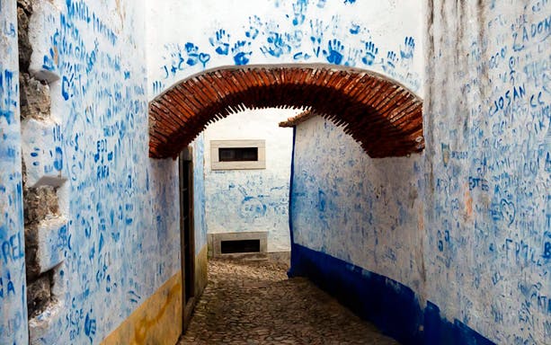 Narrow cobblestone alley in Óbidos with blue handprints and graffiti on white walls.