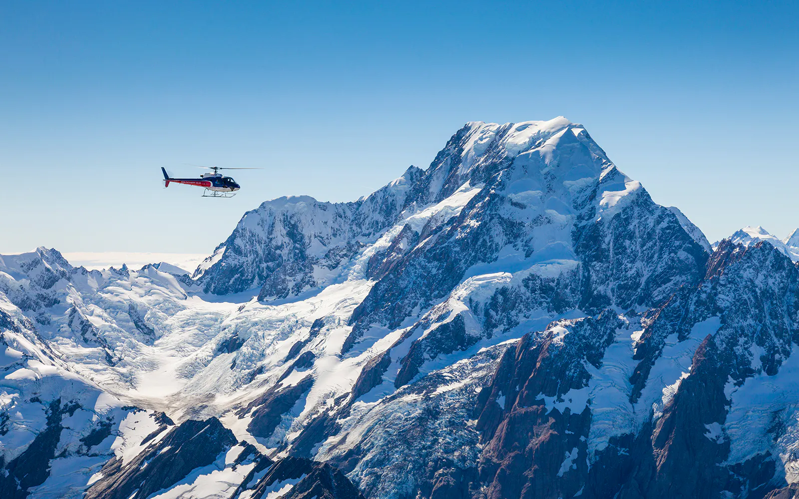 Helicopter flying near snow-covered Mount Cook, New Zealand, with alpine landing area.