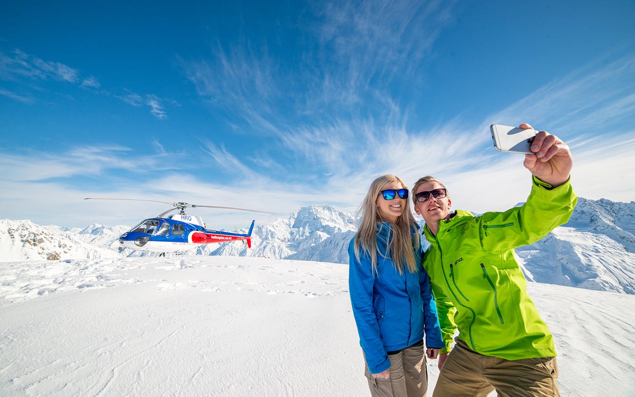 Couple taking selfie during helicopter tour on snowy Mount Cook with alpine landing.