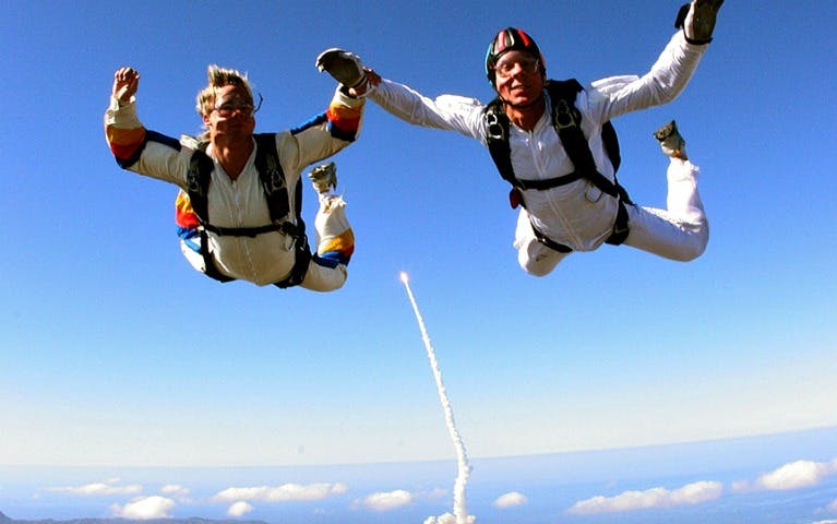 Tandem skydive over scenic landscape with rocket launch in background.