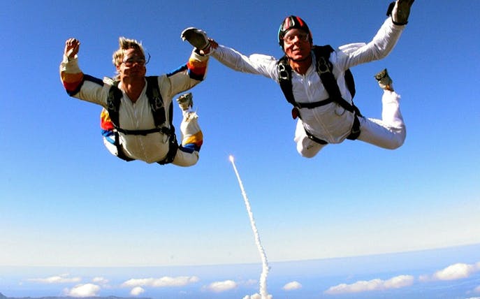 Tandem skydive over scenic landscape with rocket launch in background.