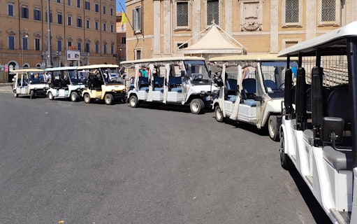 Electric golf carts lined up for a tour in Rome with historic buildings in the background.