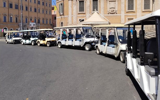 Electric golf carts lined up for a tour in Rome with historic buildings in the background.