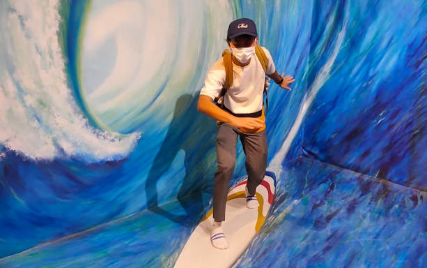 Person posing on a surfboard in 3D wave art at Illusion 3D Art Museum Melaka.