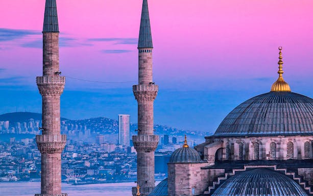 Minarets and dome of a mosque in Istanbul at sunset, with cityscape in the background.