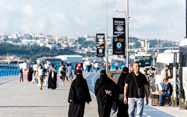 People walking along a busy promenade in Istanbul with cityscape in the background.