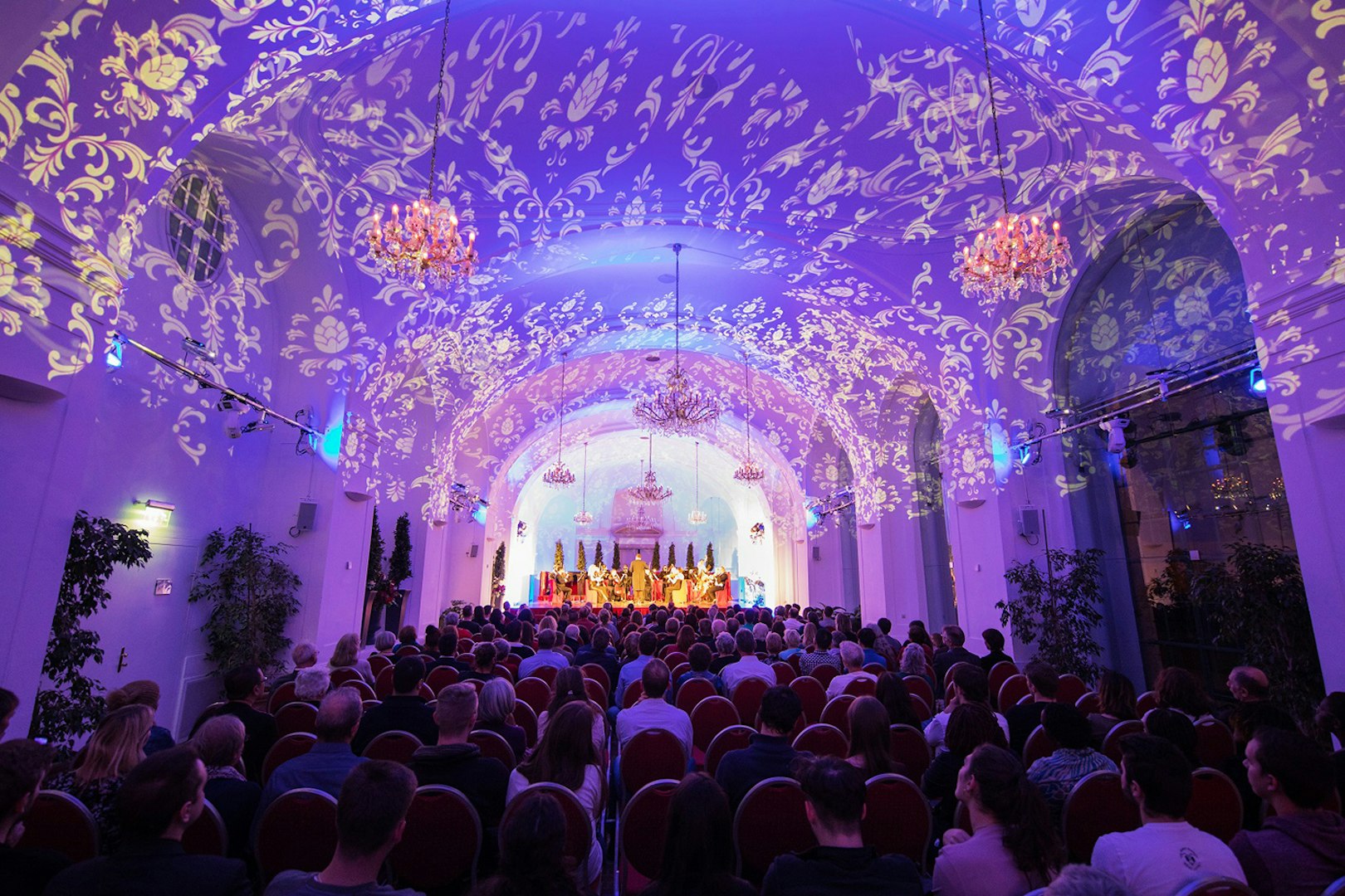 Concert audience in Schönbrunn Palace hall with decorative ceiling lights.