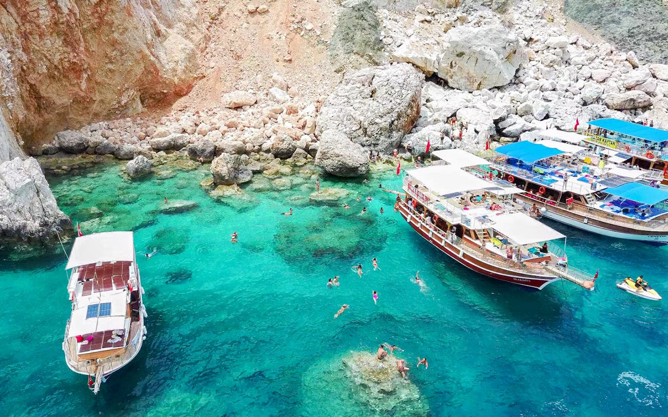 Boats anchored near rocky shore with swimmers in turquoise water, Suluada Island trip from Antalya.
