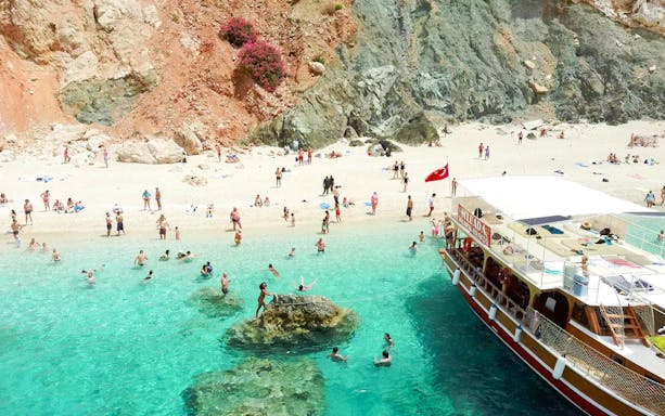 Visitors swimming near a boat at Suluada Island beach, Antalya, with rocky cliffs in the background.