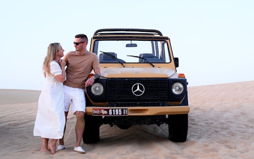 Couple leaning on vintage Mercedes in Al Marmoom Oasis desert during morning heritage safari.