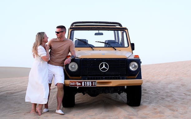 Couple leaning on vintage Mercedes in Al Marmoom Oasis desert during morning heritage safari.