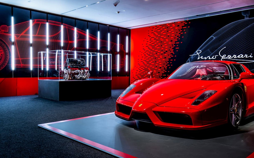 Ferrari Enzo displayed at Ferrari Museum Maranello with engine exhibit in background.