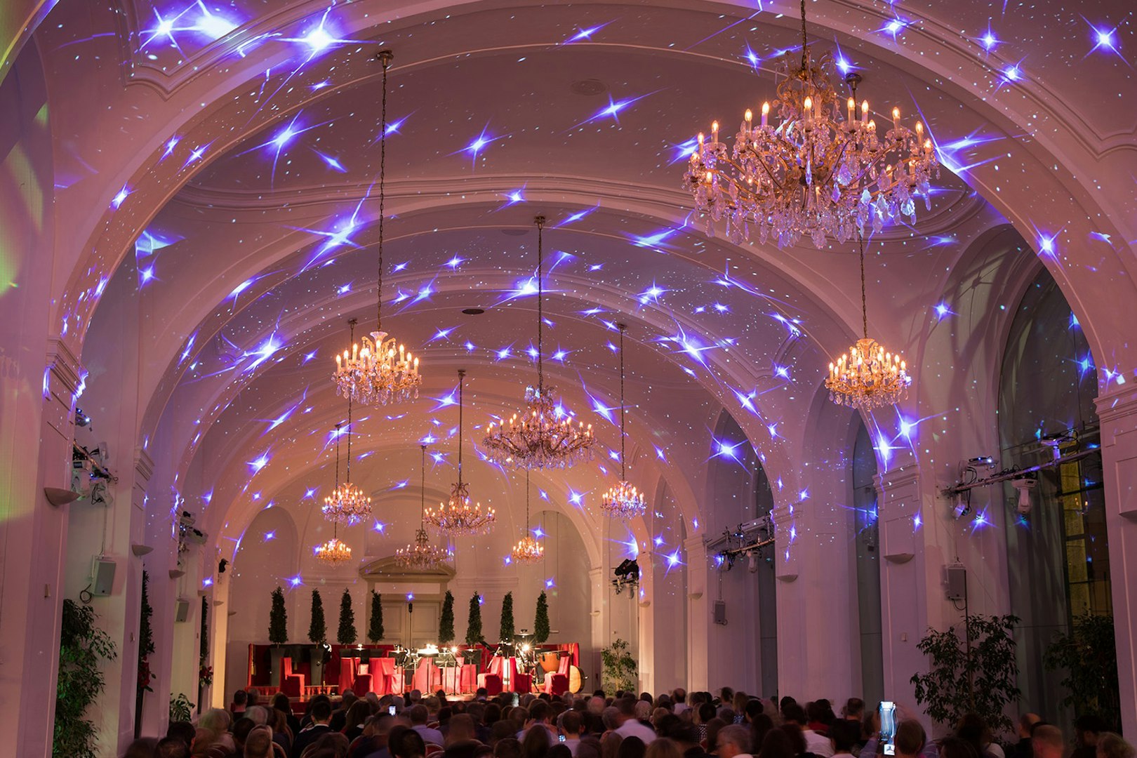 Concert audience in Schönbrunn Palace hall with chandeliers and starry light display.
