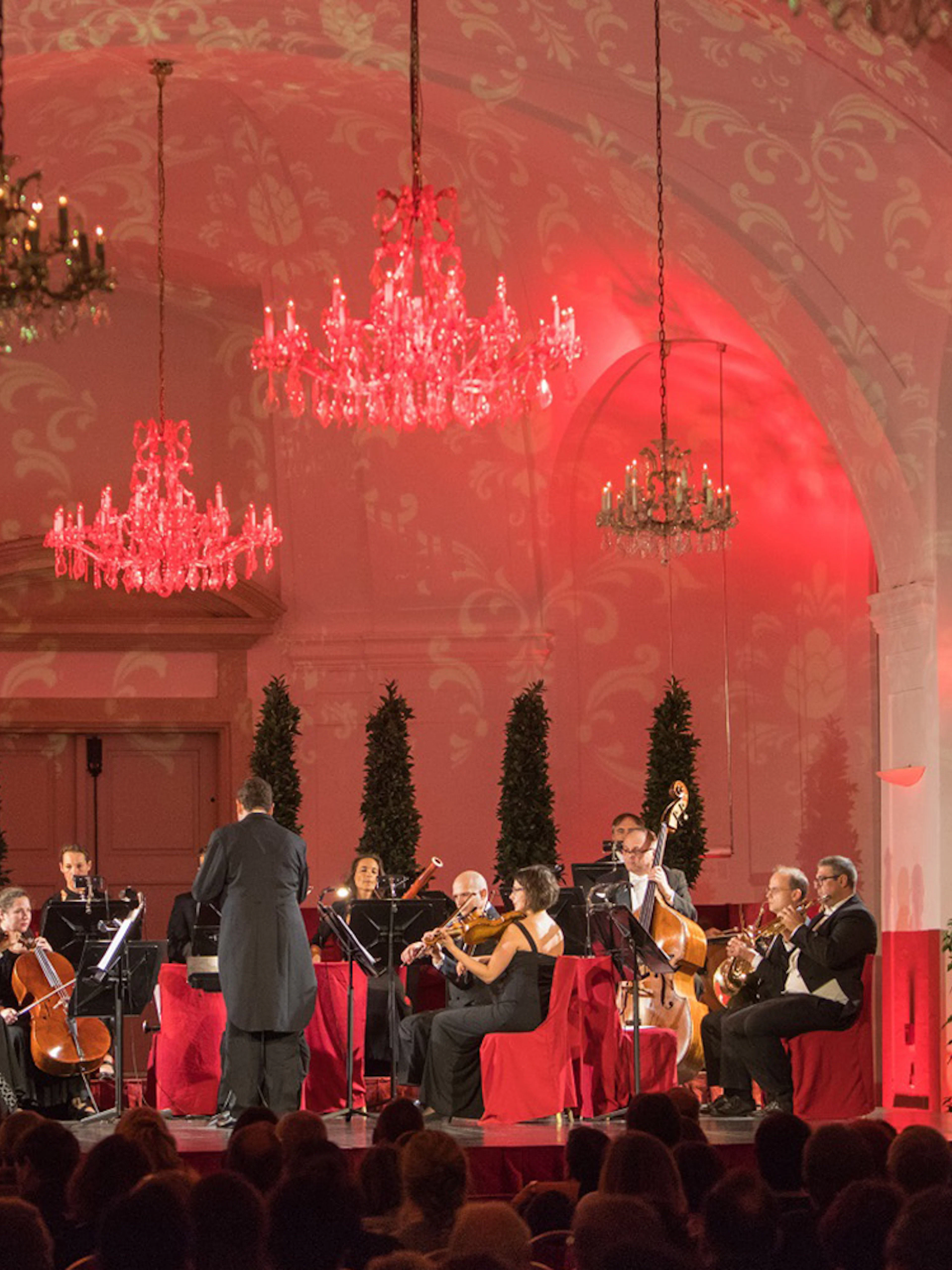 Orchestra performing at Schönbrunn Palace concert hall with chandeliers and red lighting.