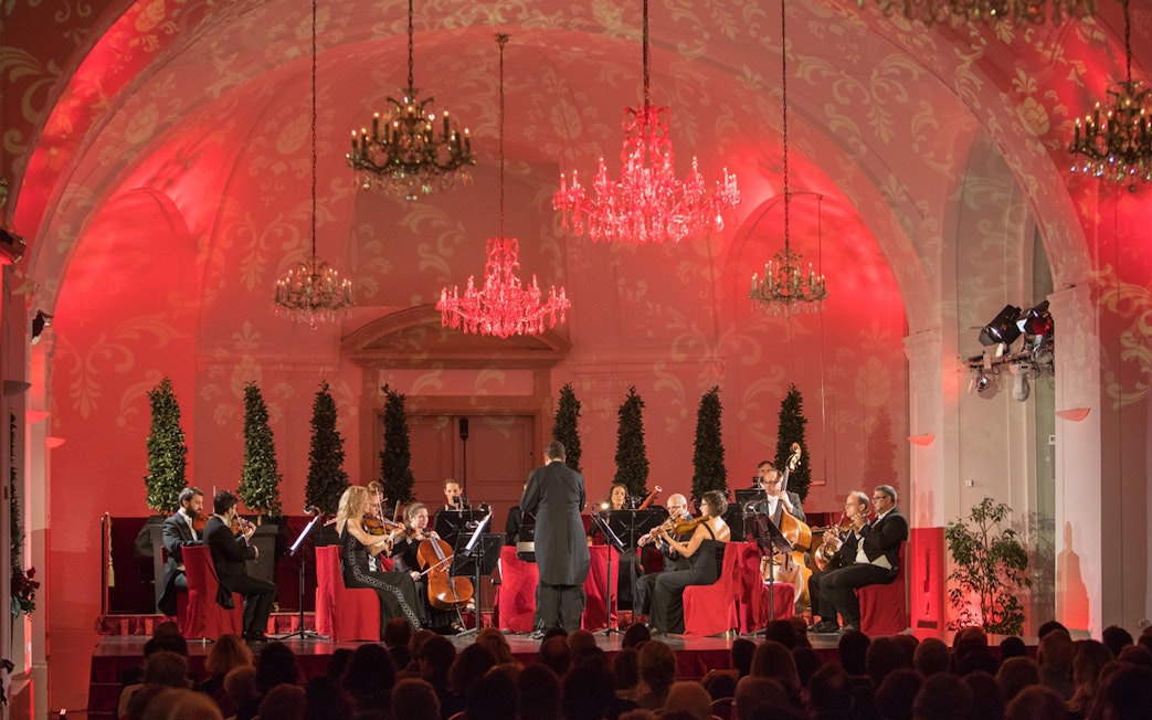 Orchestra performing at Schönbrunn Palace concert hall with chandeliers and red lighting.