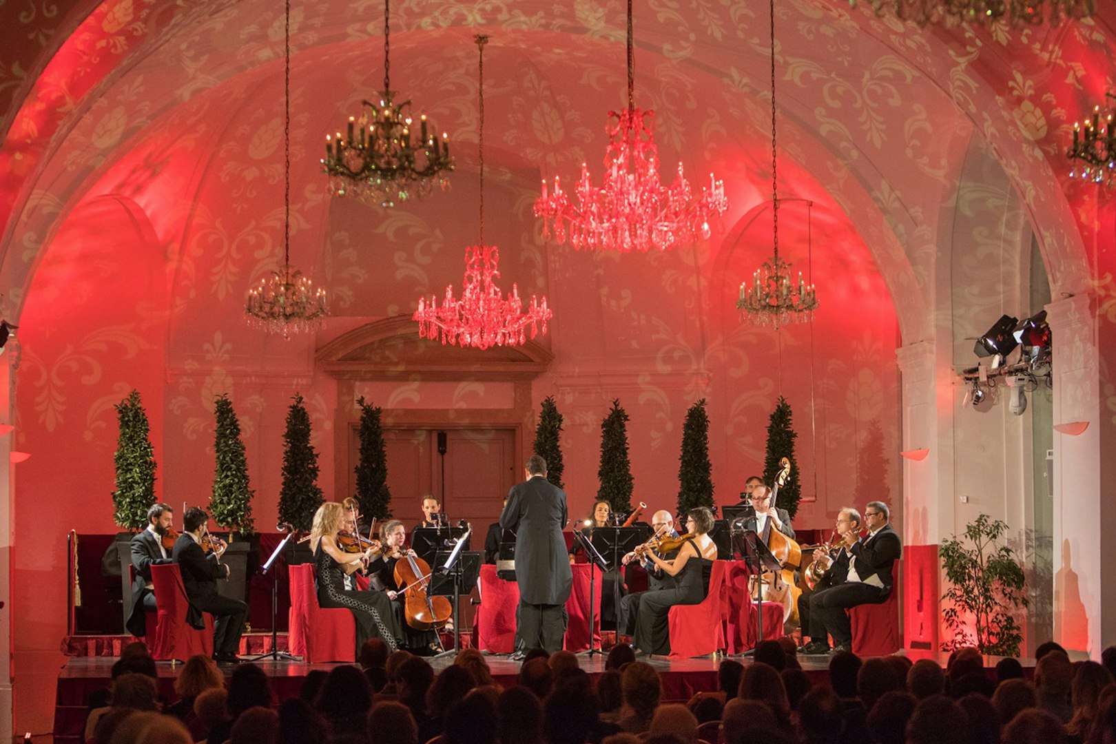 Orchestra performing at Schönbrunn Palace concert hall with chandeliers and red lighting.