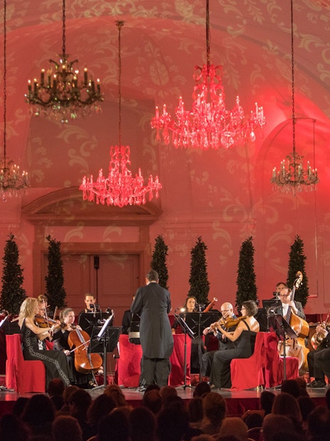 Orchestra performing at Schönbrunn Palace concert hall with chandeliers and red lighting.