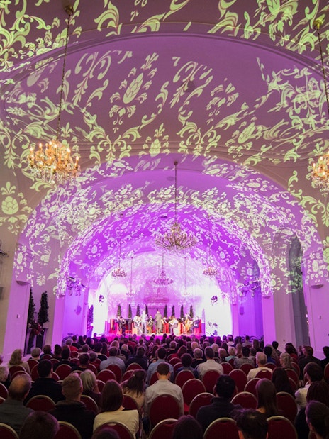 Concert audience in Schönbrunn Palace hall with ornate ceiling projections.