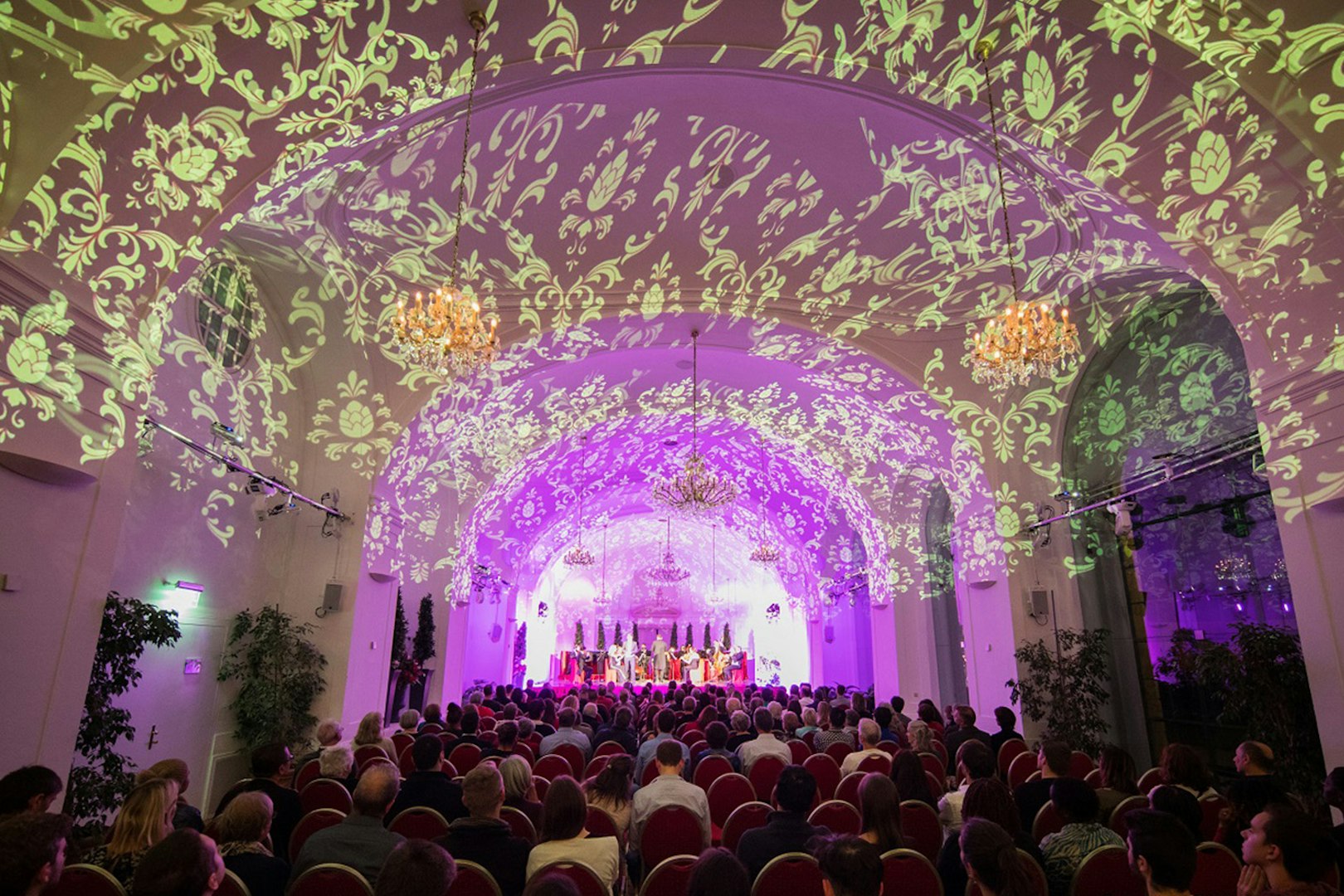 Concert audience in Schönbrunn Palace hall with ornate ceiling projections.