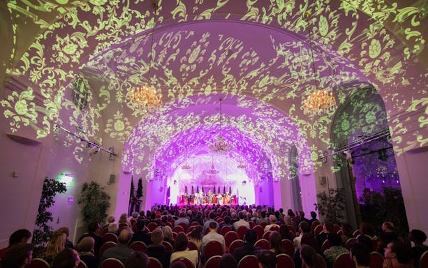 Concert audience in Schönbrunn Palace hall with ornate ceiling projections.
