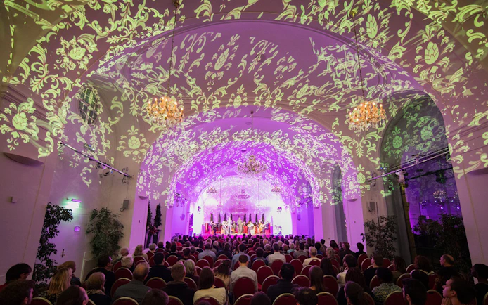 Concert audience in Schönbrunn Palace with ornate ceiling projections during Mozart & Strauss performance.