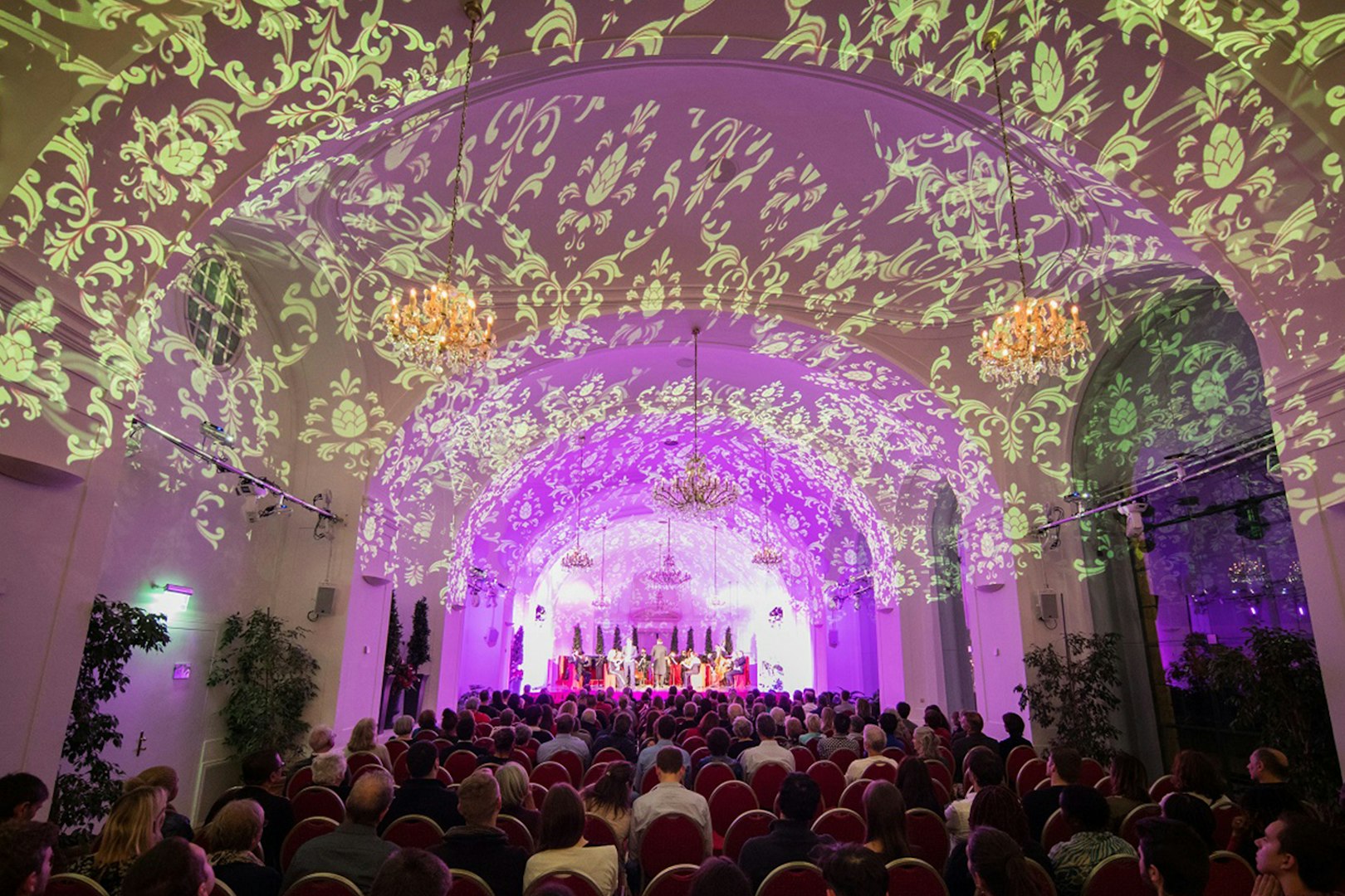 Concert audience in Schönbrunn Palace with ornate ceiling projections during Mozart & Strauss performance.