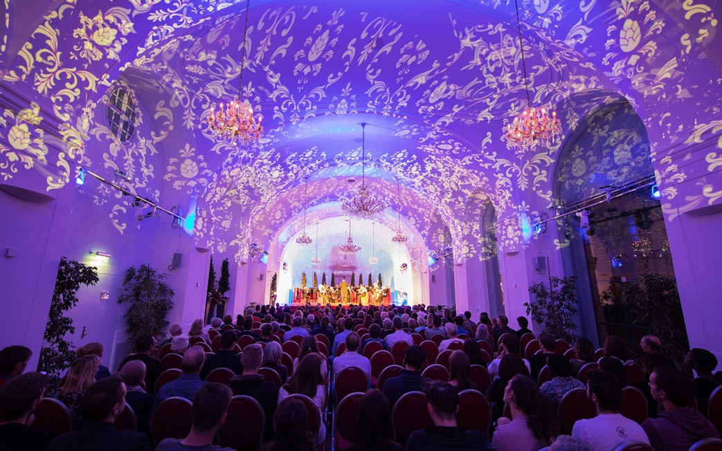 Concert audience in Schönbrunn Palace hall with ornate ceiling projections.