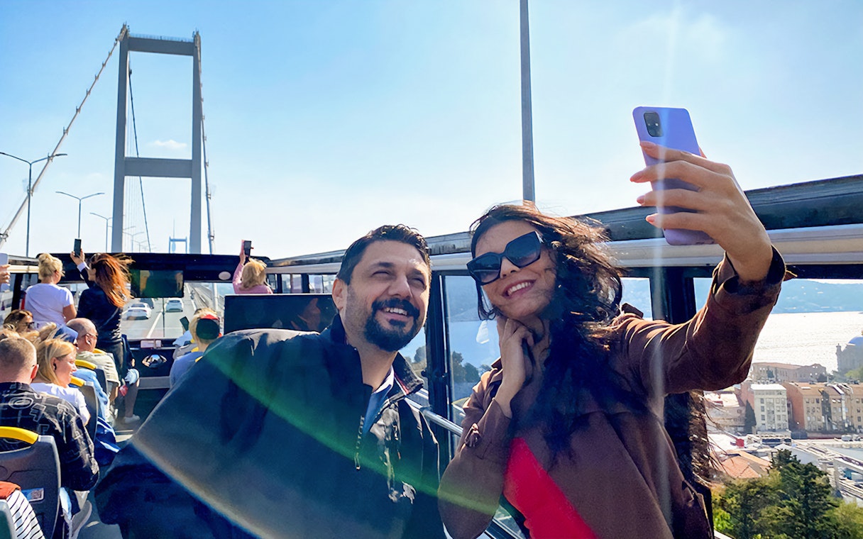 Tourists on Istanbul hop-on hop-off bus with Bosphorus Bridge in background.