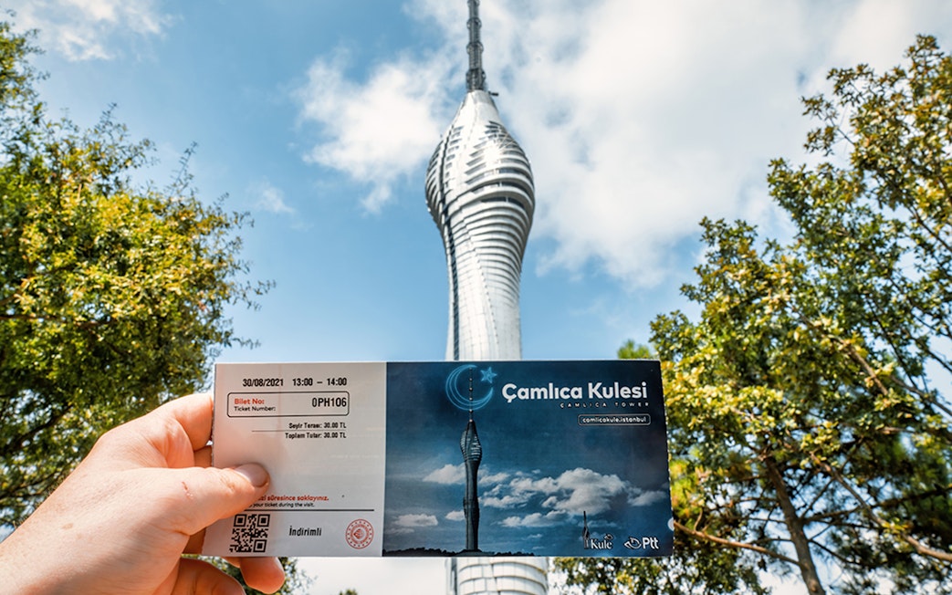 Hand holding a ticket in front of Çamlıca Istanbul TV Tower against a blue sky.