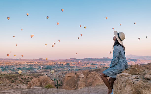 Hot air balloons over Cappadocia at sunrise, woman watching from rocky viewpoint.