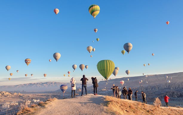 Hot air balloons over Cappadocia at sunrise with tourists watching from a hilltop.