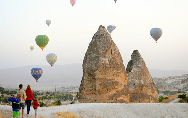 Hot air balloons over Cappadocia's rock formations at sunrise, with a family watching.