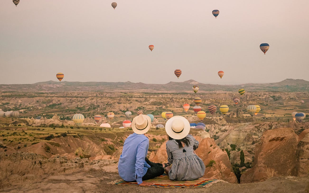 Couple watching hot air balloons over Cappadocia landscape at sunrise.