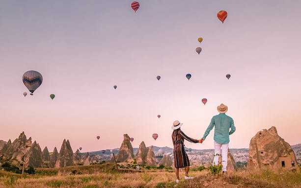 Couple watching hot air balloons over Cappadocia's rock formations at sunrise.