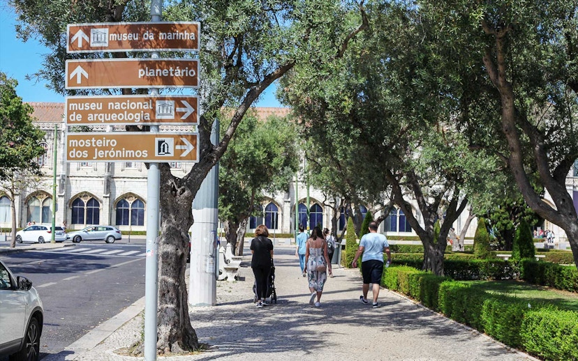 Signage for Jerónimos Monastery and Navy Planetarium in Lisbon, with people walking nearby.