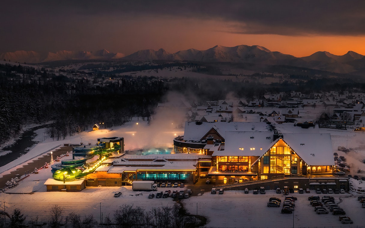 Chocholow Thermal Baths illuminated at dusk with snowy Tatra Mountains in the background.