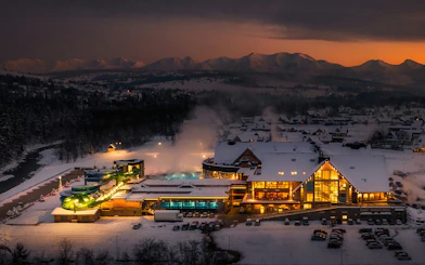 Chocholow Thermal Baths illuminated at dusk with snowy Tatra Mountains in the background.