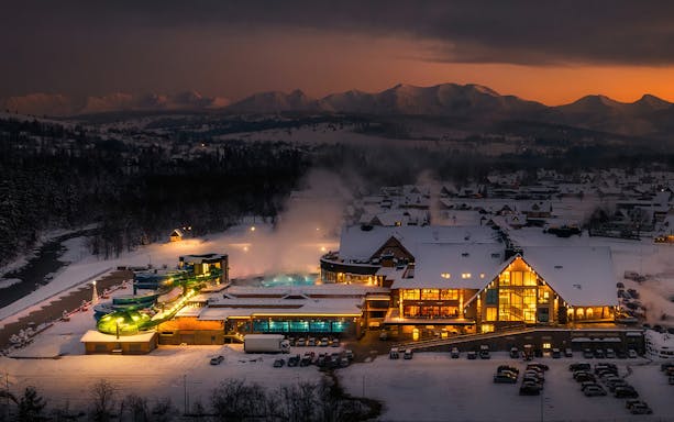 Chocholow Thermal Baths illuminated at dusk with snowy Tatra Mountains in the background.
