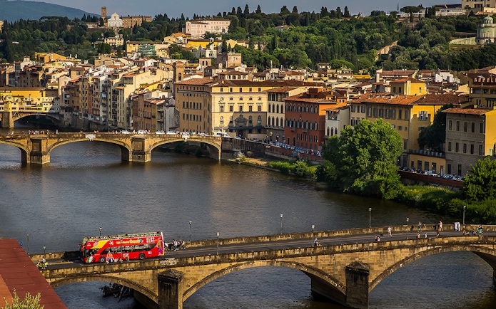 Open-top tour bus crossing a bridge in Florence with cityscape in the background.