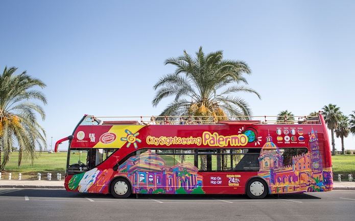 Palermo hop-on hop-off bus with colorful cityscape design parked by palm trees.