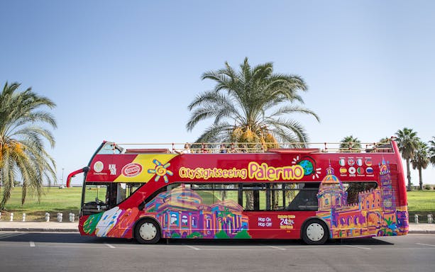 Palermo hop-on hop-off bus with colorful cityscape design parked by palm trees.